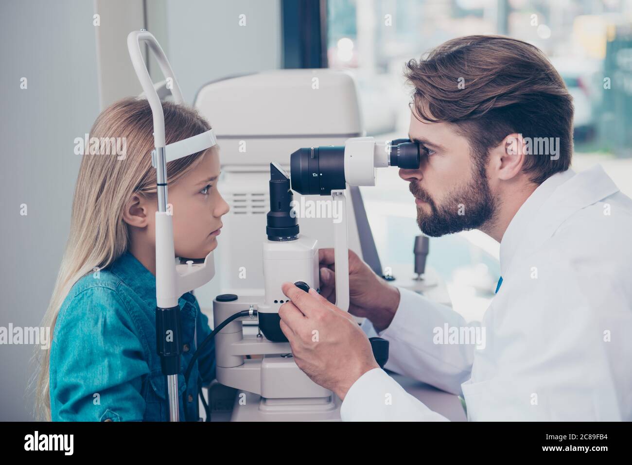 Health care, medicine, eye sight and technology concept. Focused brunet bearded optometrist with non contact tonometer is checking small blond`s girl Stock Photo